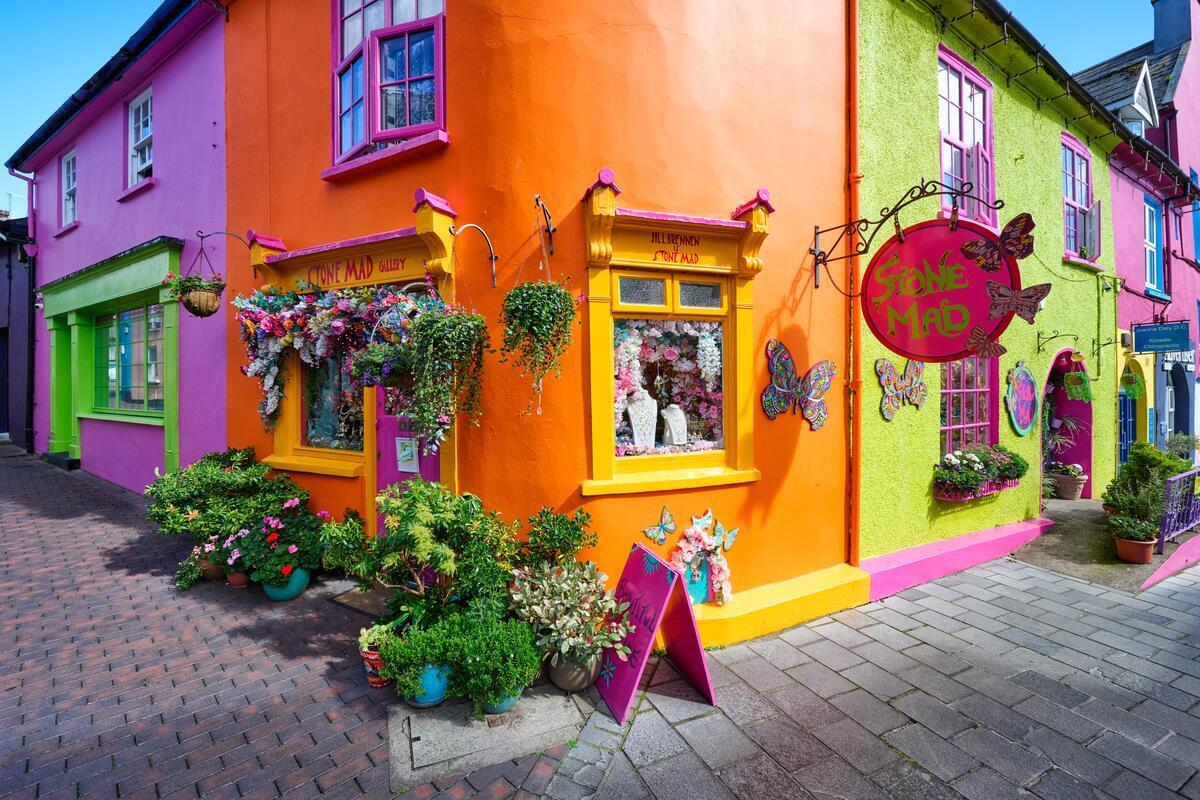 A brightly painted street corner in Kinsale, County Cork, with orange, pink and green shopfronts, flowers and colourful signage.